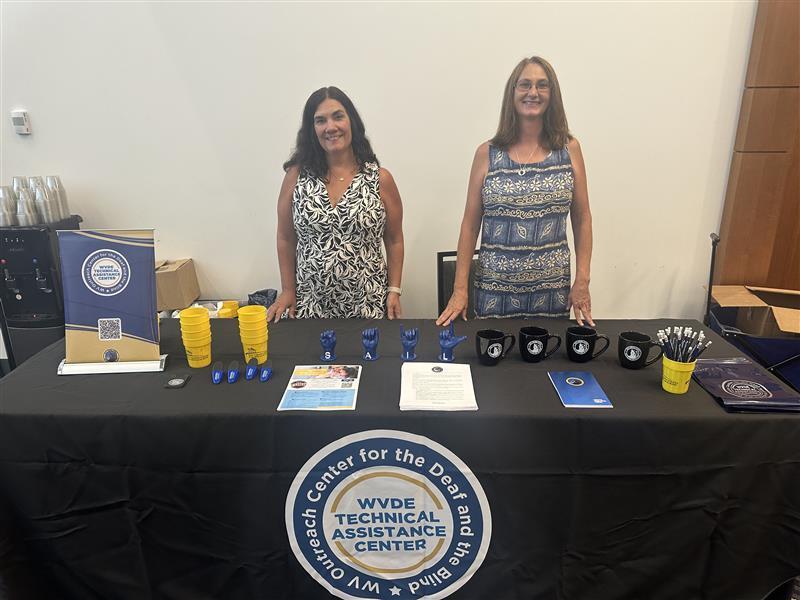Two females, Mrs. Nora Stevenson and Ms. Wendy Bailey stand behind a table that is covered with a black tablecloth. On top of the table is a variety of giveaway items and informational packets. 