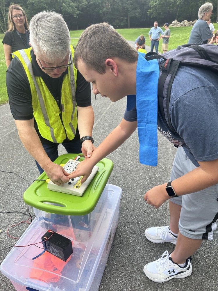 A male scientist wearing a bright yellow vest assists a male student with pressing the button to launch his rocket. 