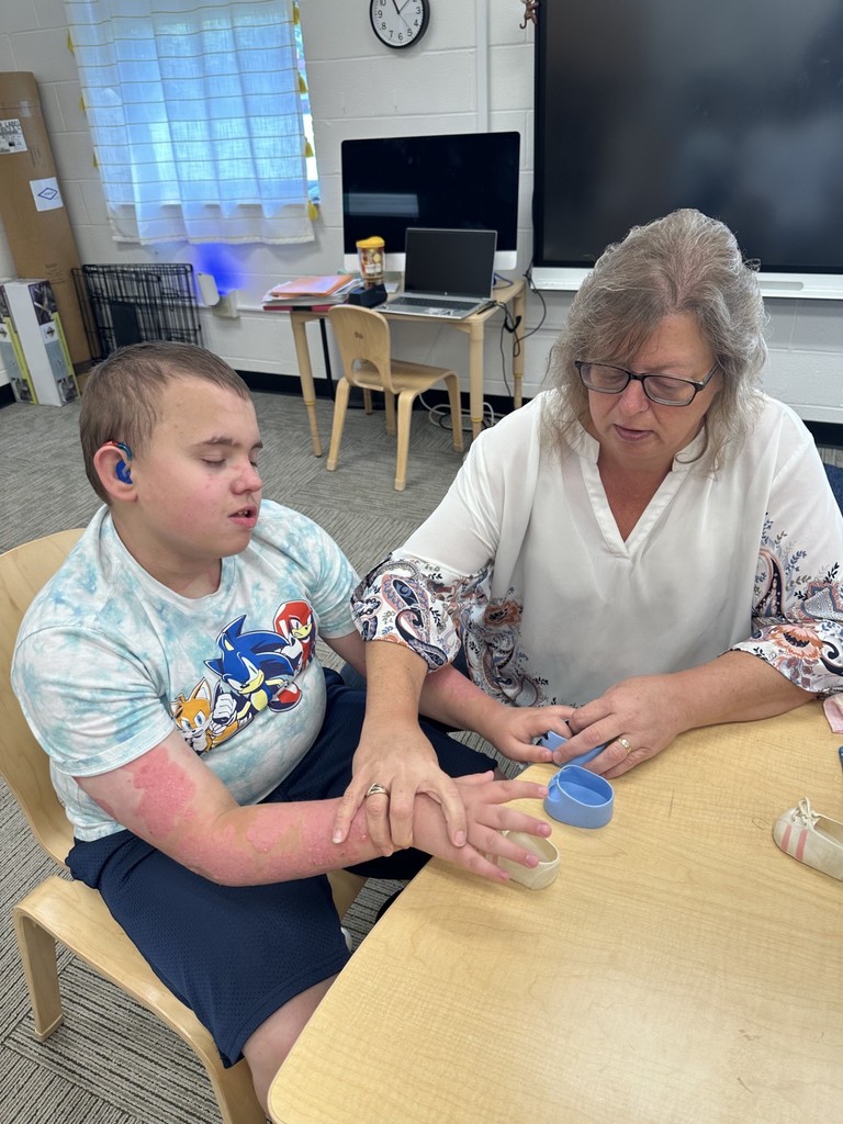 A female intervenor looks down as she assists a male student with picking up objects off of a table.