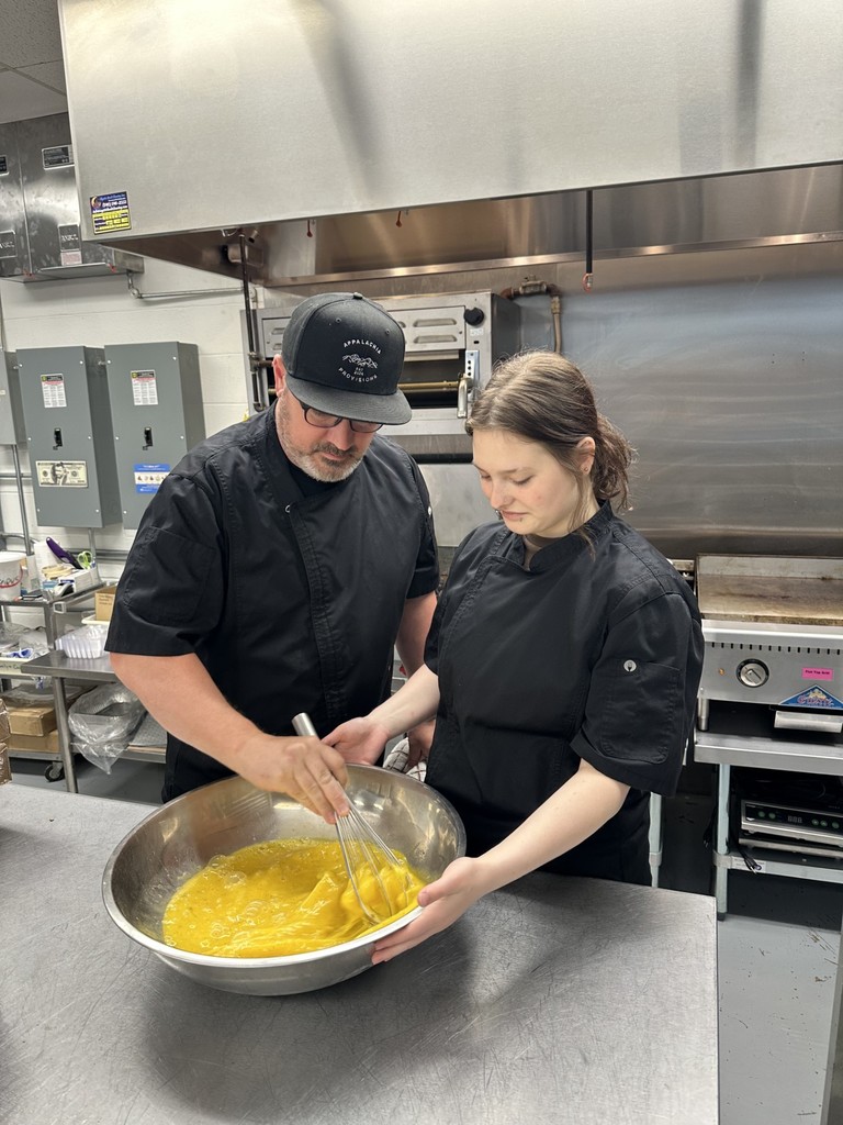 A male culinary instructor assists a student with whisking eggs in a large metal bowl.