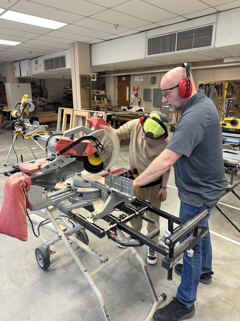 A male instructor assists a male student with using a table saw.