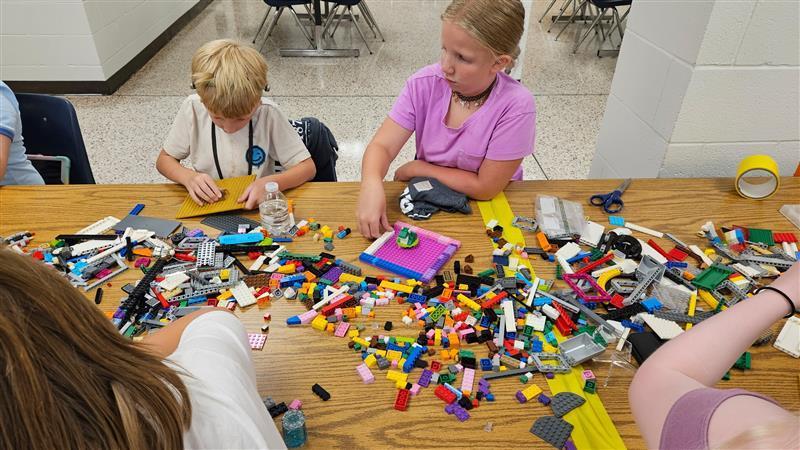 Students seated at a rectangular-shaped table while different colored LEGO pieces scatter across the table.