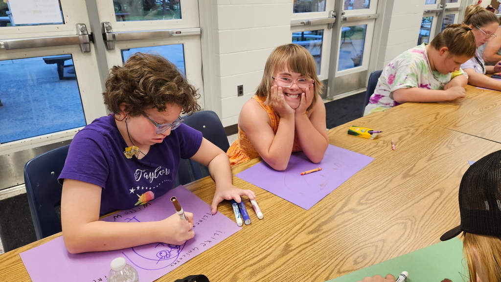 Two female students seated at a rectangular-shaped table. One female looks down as she draws on a piece of paper, while the other female smiles at the camera.