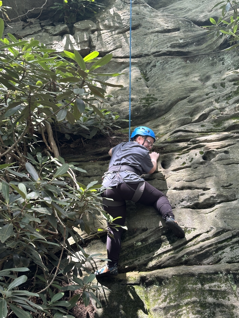 A female camp participant wearing a blue rock climbing helmet climbs up a wall.