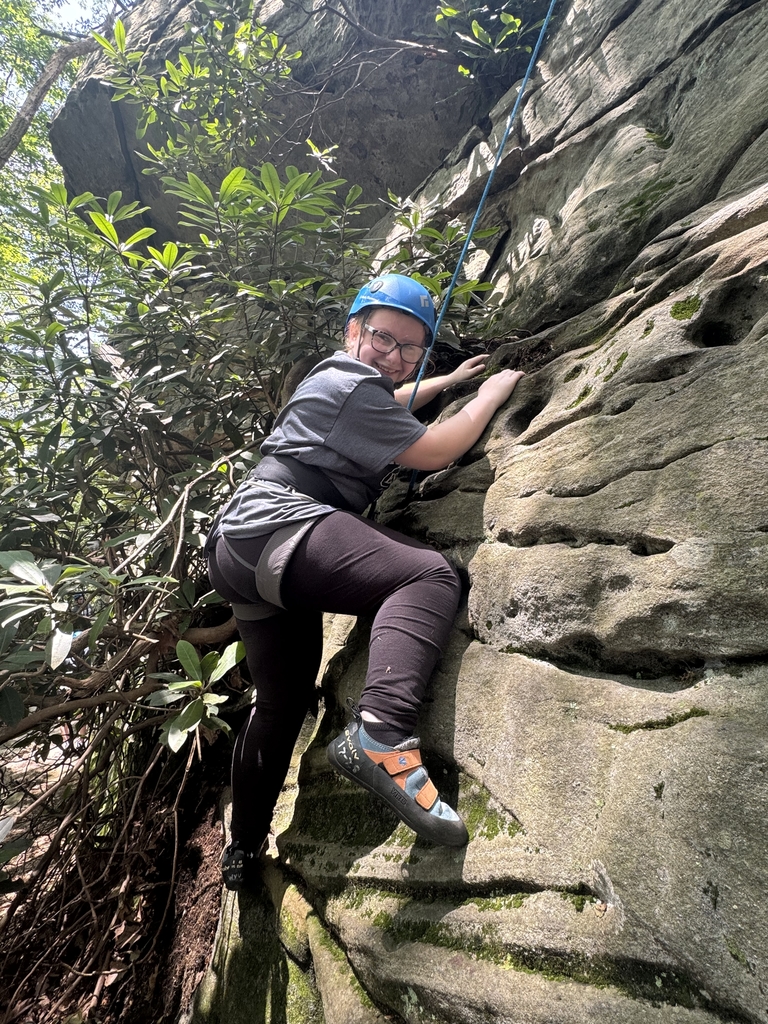 A female camp participant looks down and smiles for the camera as she climbs up the side of a large rock.