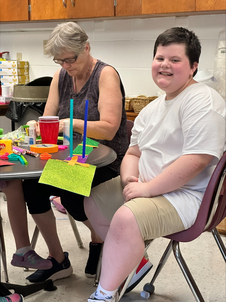 A male camp participant seated at a circular-shaped table smiles at the camera with his bridge building materials in front of him.