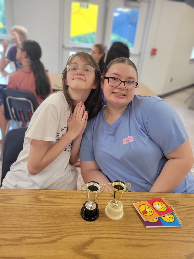 Two female camp participants are seated at a table side-by-side while two trophies sit on the table in front of them. 