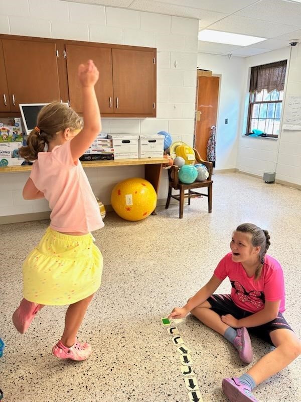 A female camp participant jumps into the air excitedly as another female camp participant who is seated on the ground looks up at her and smiles.