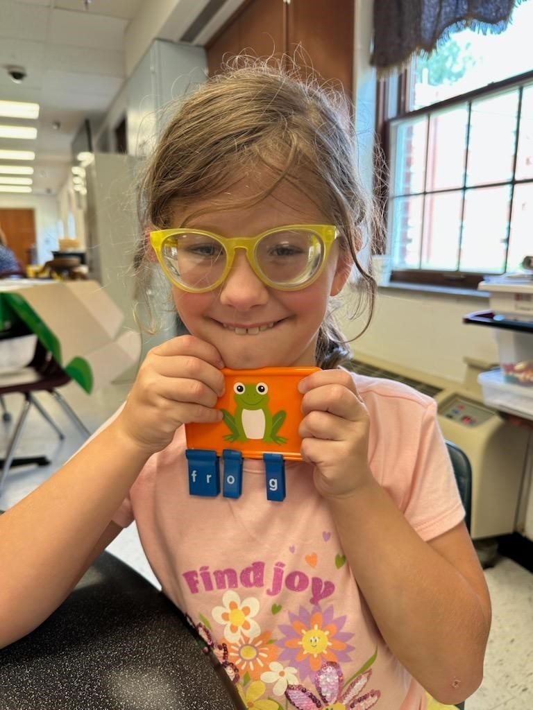 A female camp participant wearing yellow glasses smiles at the camera as she holds up a frog with the letters "F,R,O,G' spelling out the word. 