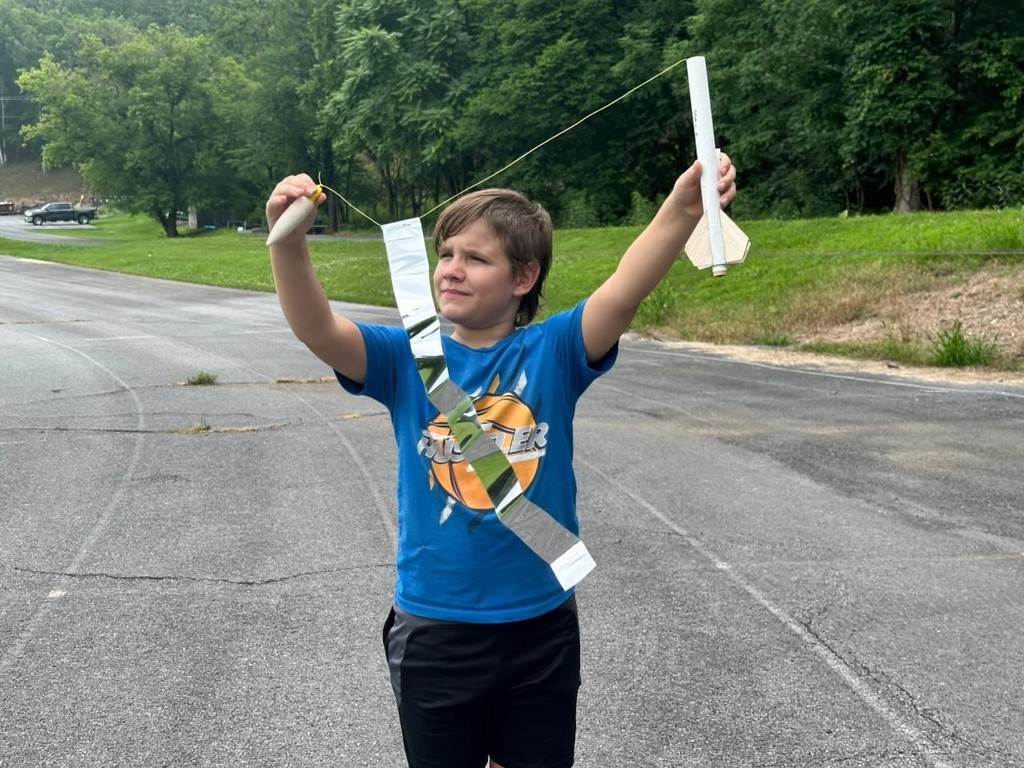 A male camp participant wearing a blue shirt holds up a wooden rocket that was launched into the sky. 