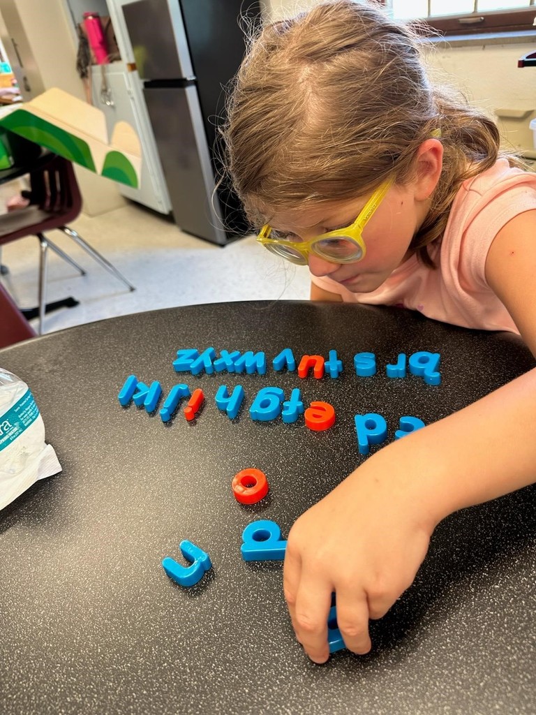 A female camp participant wearing yellow glasses looks down as she places blue and red letters into a sentence. 
