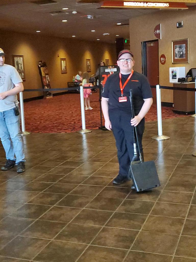 A male individual wearing an all-black work uniform, smiles for the camera as he holds a broom in his left hand.