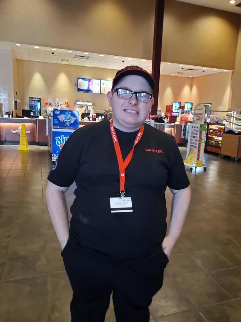 Cody Dean. A male individual stands proudly with his hands in his pockets while smiling at the camera. A movie theatre snack bar is in the background.