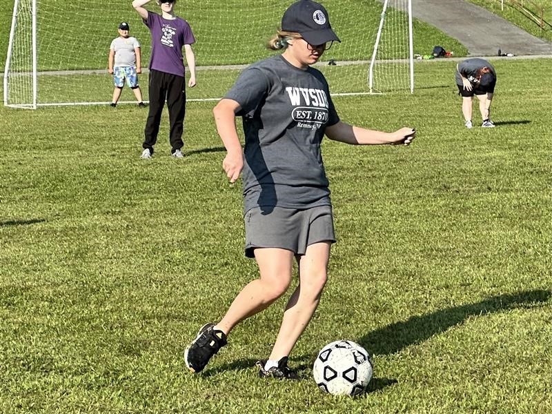 A female camp participant wearing a gray-colored shirt and navy-colored hat runs as she dribbles a black and white-colored soccer ball. 