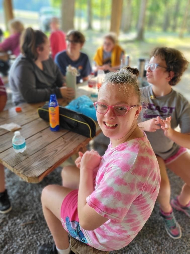 A female camp participant wearing a pink and white tie-dye shirt and pink glasses smiles at the camera as she is seated at a wooden picnic table. 