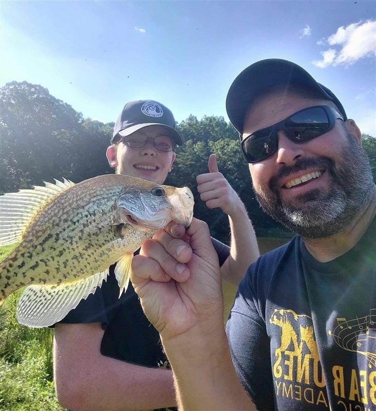 A male teacher wearing a hat and sunglasses poses with a male student who is wearing a hat and glasses pose with a fish.