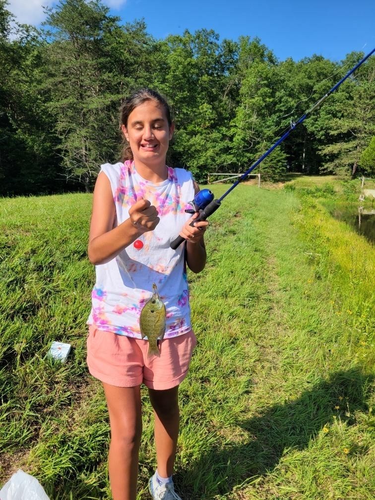 A female camp participant wearing a tie-dyed shirt smiles for the camera as she holds a blue fishing rod in one hand and a fish in the other. 