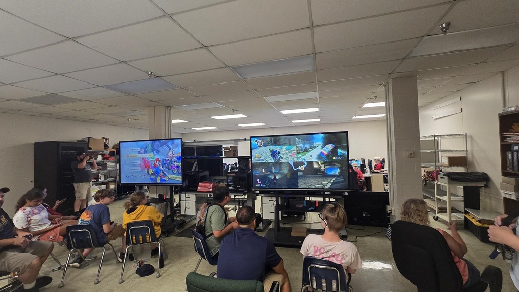 Camp participants seated in chairs as they watch large screens displaying video games. 