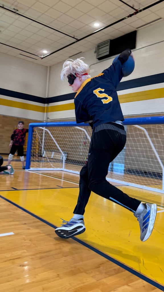 A male camp participant wearing a blue and gold-colored jersey with the number "5" on the back gets ready to roll a blue goalball.