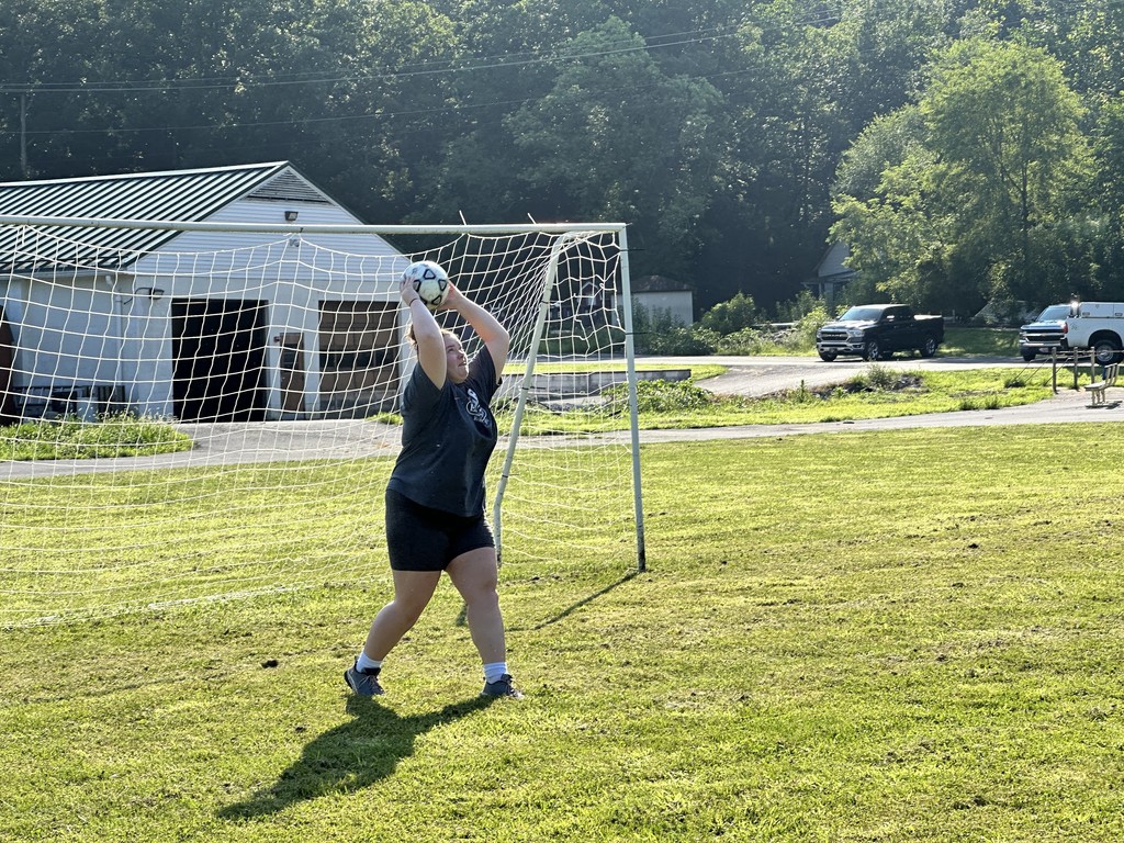 A female camp participant is standing in the soccer goal net as she throws in the black and white-colored soccer ball. 