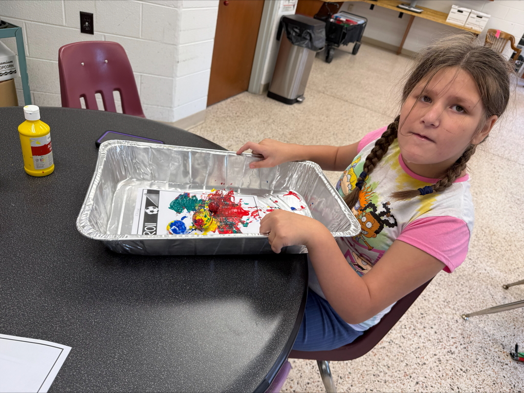 A female camp participant is seated at a circular table as she looks at the camera while working on her paint project.