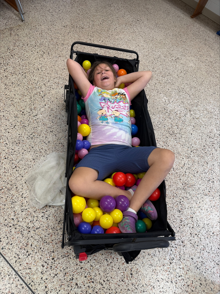 A female camp participant wearing a tie-dye shirt is laying in a cart full of different colored balls. 
