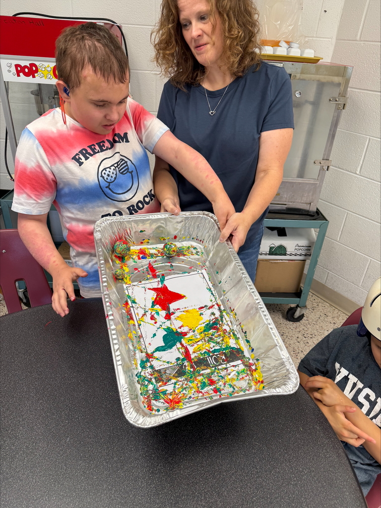 A male camp participant wearing a tie-dye shirt is assisted by a female teacher wearing a navy-colored shirt with painting.