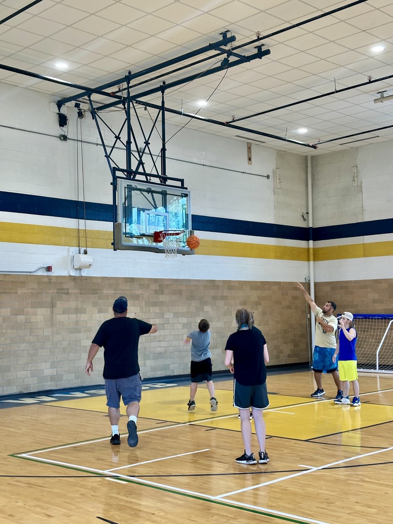 Camp participants are gathered around one another as they shoot an orange basketball into the hoop inside of a gymnasium. 