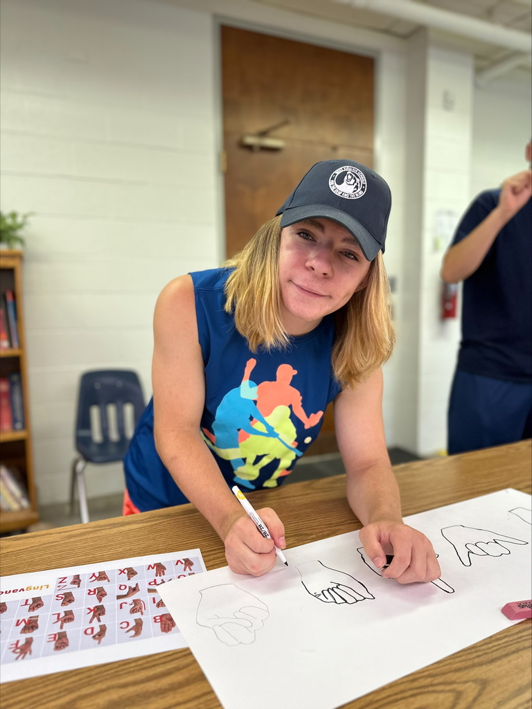 A male camp participant wearing a blue shirt and hat holds a marker while he looks up at the camera and smiles. 