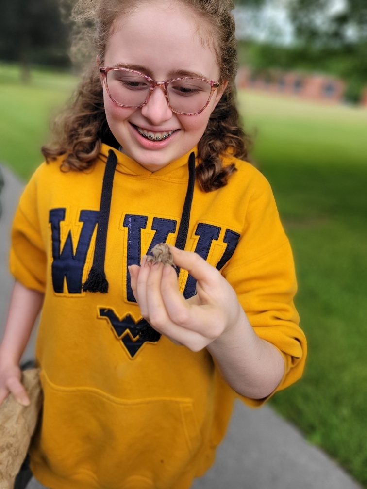 A female camp participant wearing a bright yellow sweatshirt and curly blonde hair smiles as she holds an insect in her hand.