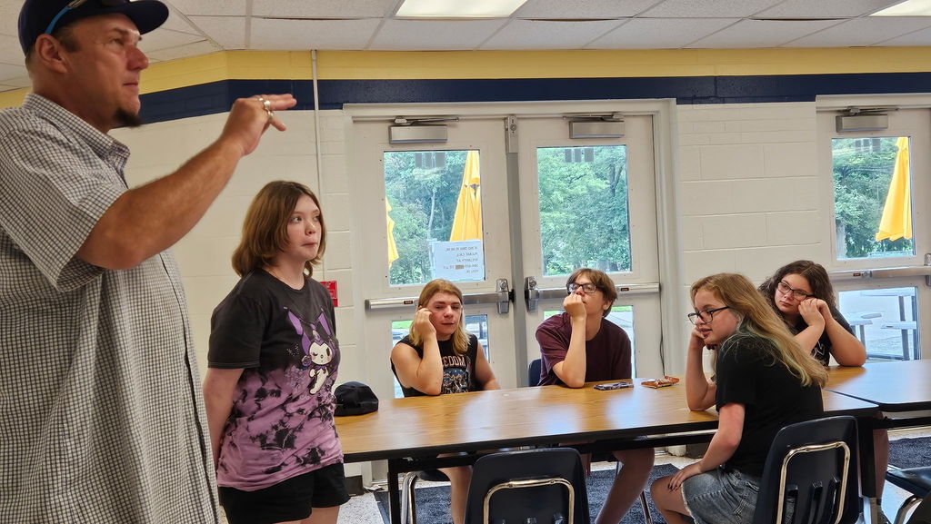 A male residential care specialist uses ASL to communicate with camp participants. There is a female camp participant standing to his left. There are campers seated at a table in the background. 