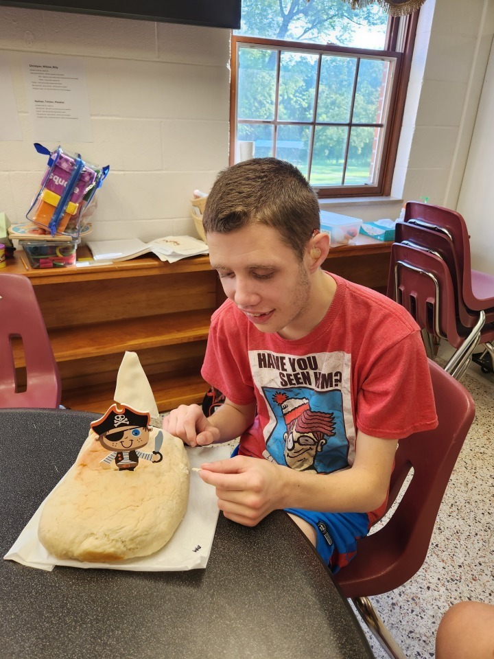 A male camp participant looks down and smiles at his pirate ship shaped bread creation. 