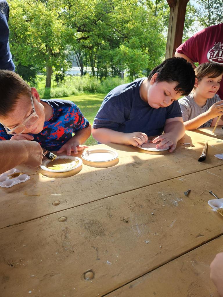 Three male camp participants are seated at a wooden picnic table as they paint white-colored Styrofoam plates the color "gold."