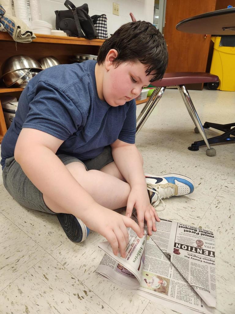 A male camp participant sits on the ground with his legs crossed. He looks down and concentrates on making his newspaper pirate hat. 