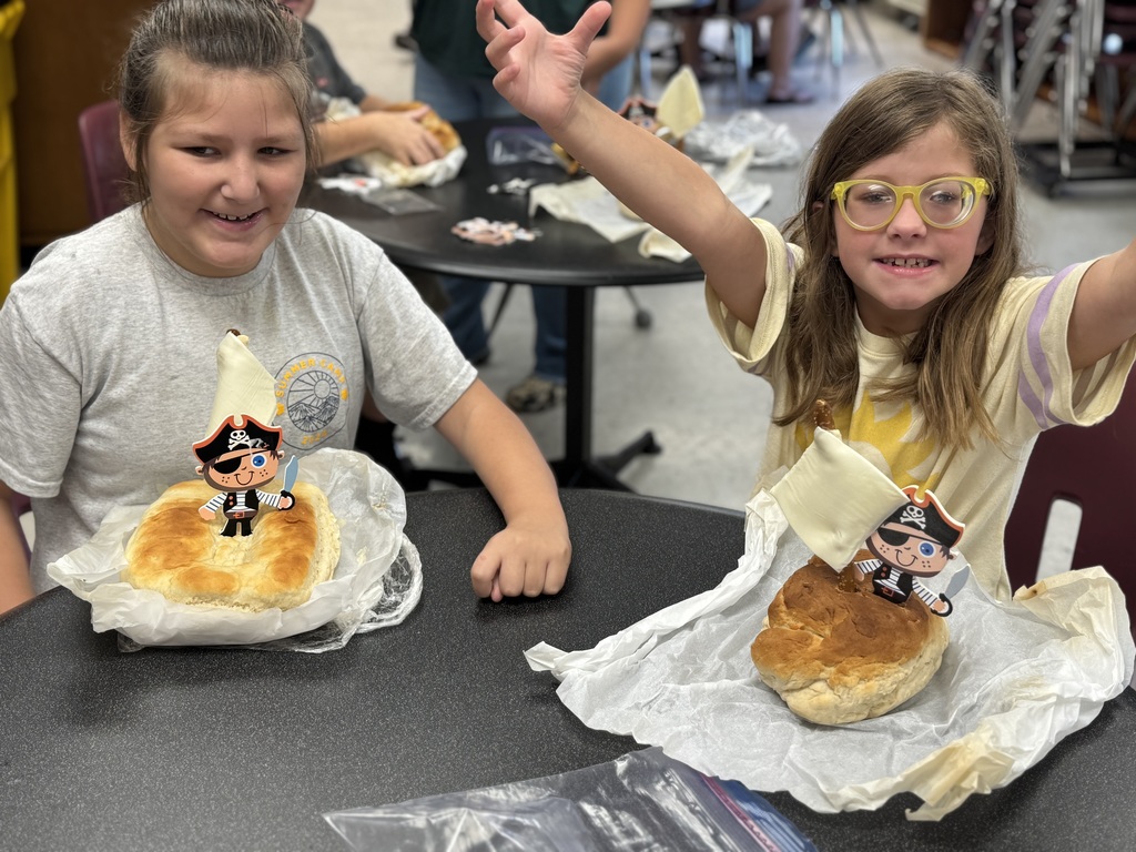 Two female camp participants seated at a circular shaped table pose excitedly with their pirate ship shaped bread. 