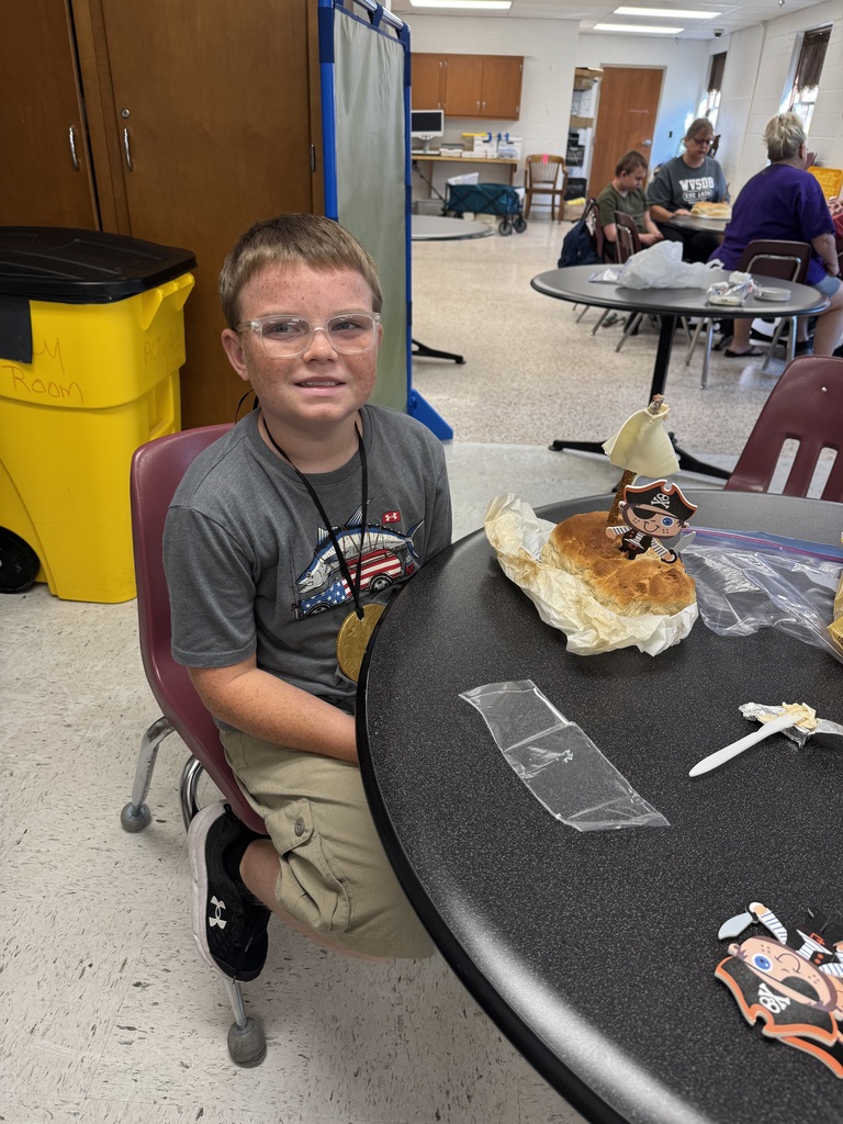 A male camp participant seated at a circular shaped table smiles for the camera with his pirate ship shaped bread. 