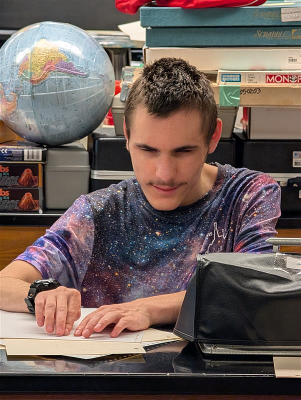 A male camp participant wearing a galaxy-themed shirt touches a piece of paper that is embossed with Braille. 