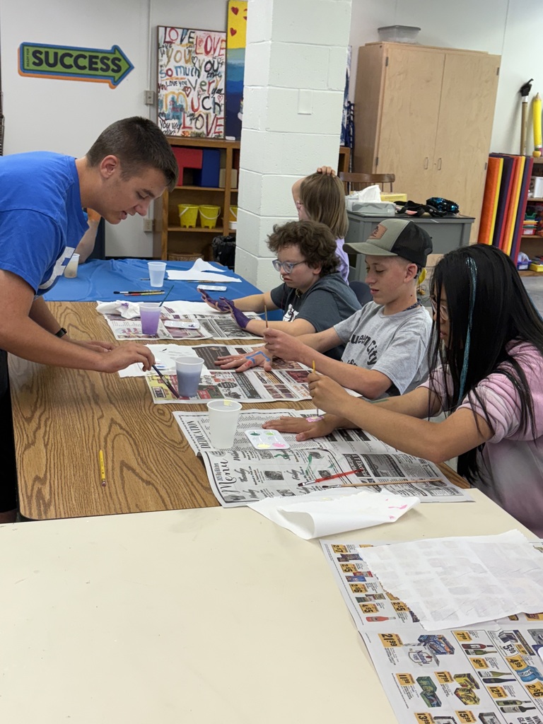 A male camp volunteer assists camp participants in an art project where they are painting on their hands. 