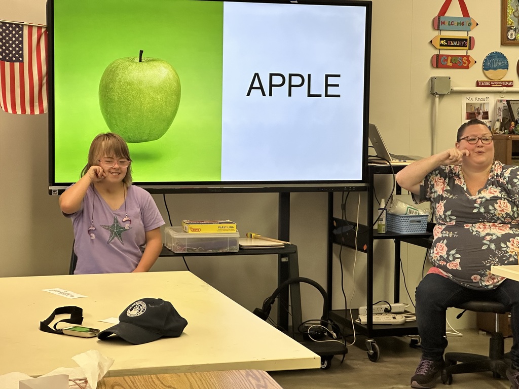 A female camp participant and female ASL teacher sign the word "apple." There is a large green-colored apple on the smartboard behind them with the word "apple" in black-font beside of the apple.