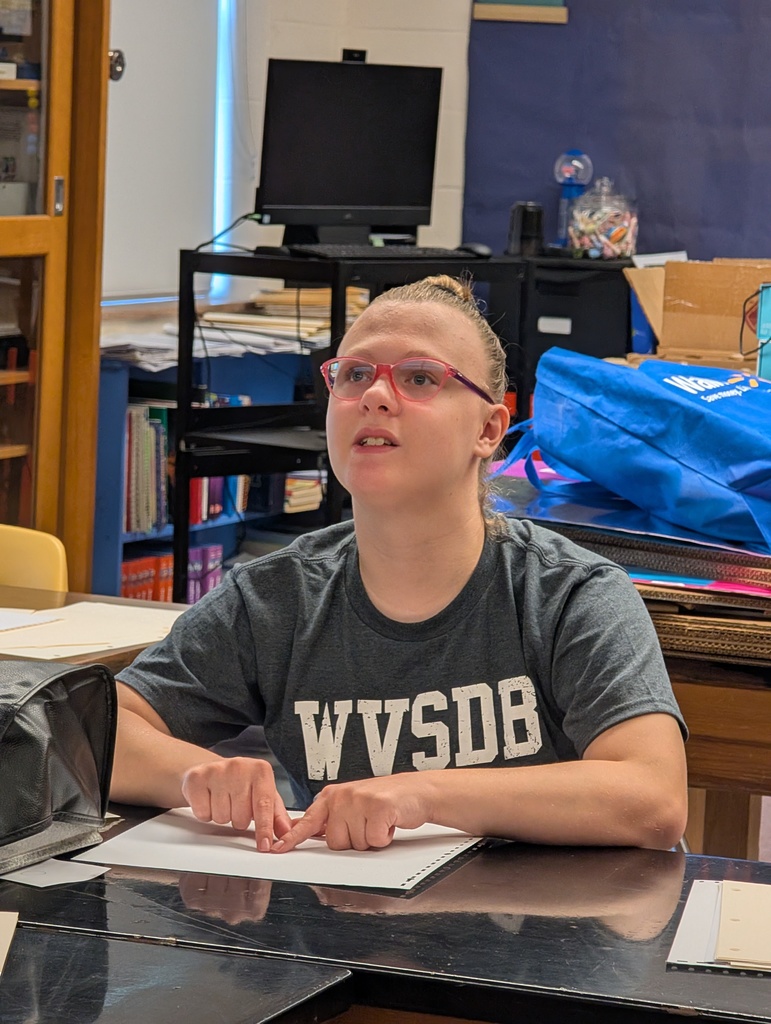 A female camp participant wearing a dark gray-colored shirt and pink-colored glasses touches a white piece of paper that is embossed with Braille.