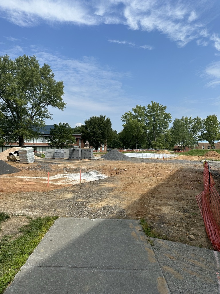 Construction site of the new greenhouse. Dirt and piles of gravel are spread out. There is a concrete slab in the background where the greenhouse will be.