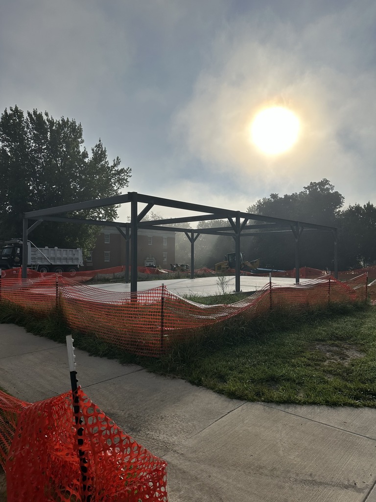 Steel beams make up the pavilion. Orange construction tape surrounds the perimeter of the pavilion. Keller Hall is in the background.