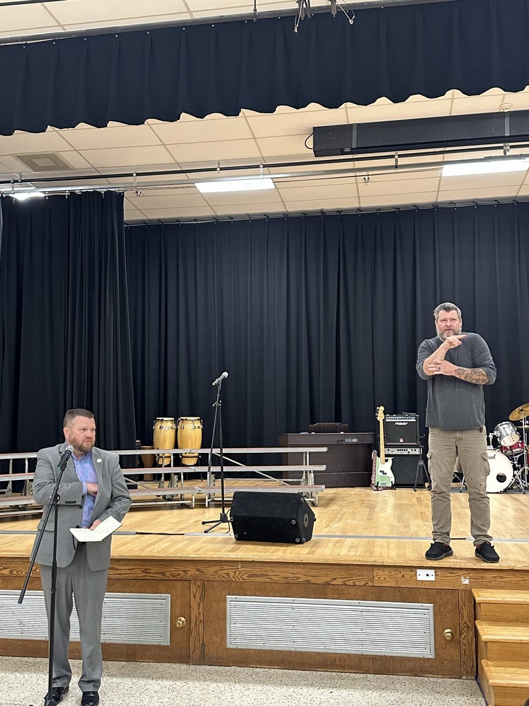Superintendent Burch stands behind a microphone as he delivers opening remarks. A male is standing on the stage behind him interpreting for him.