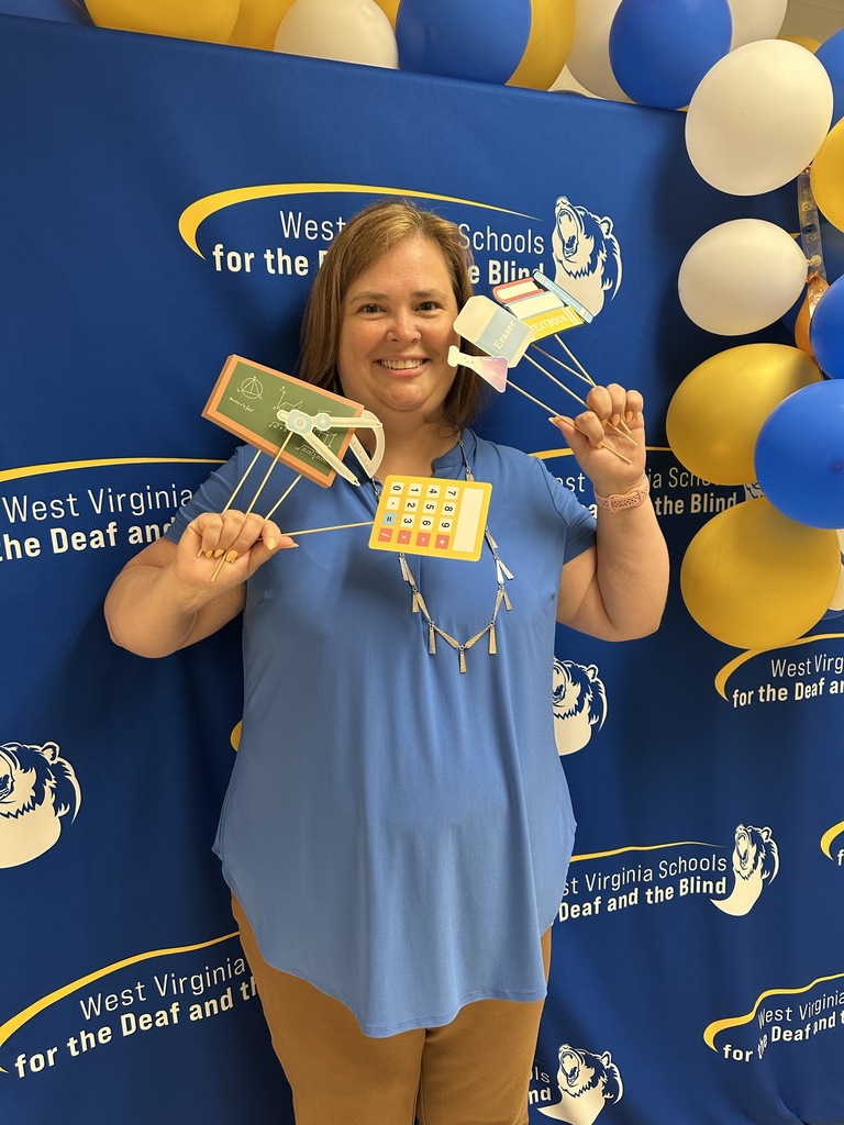 A female administrator stands in front of a blue backdrop holding many photobooth props in her hands as she smiles for the camera.
