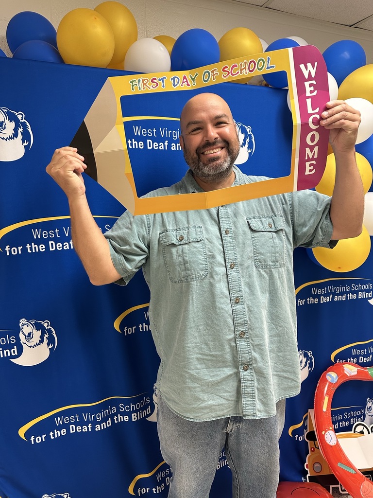 A male individual stands in front of a blue backdrop holding a pencil photobooth backdrop that says "First Day of School."
