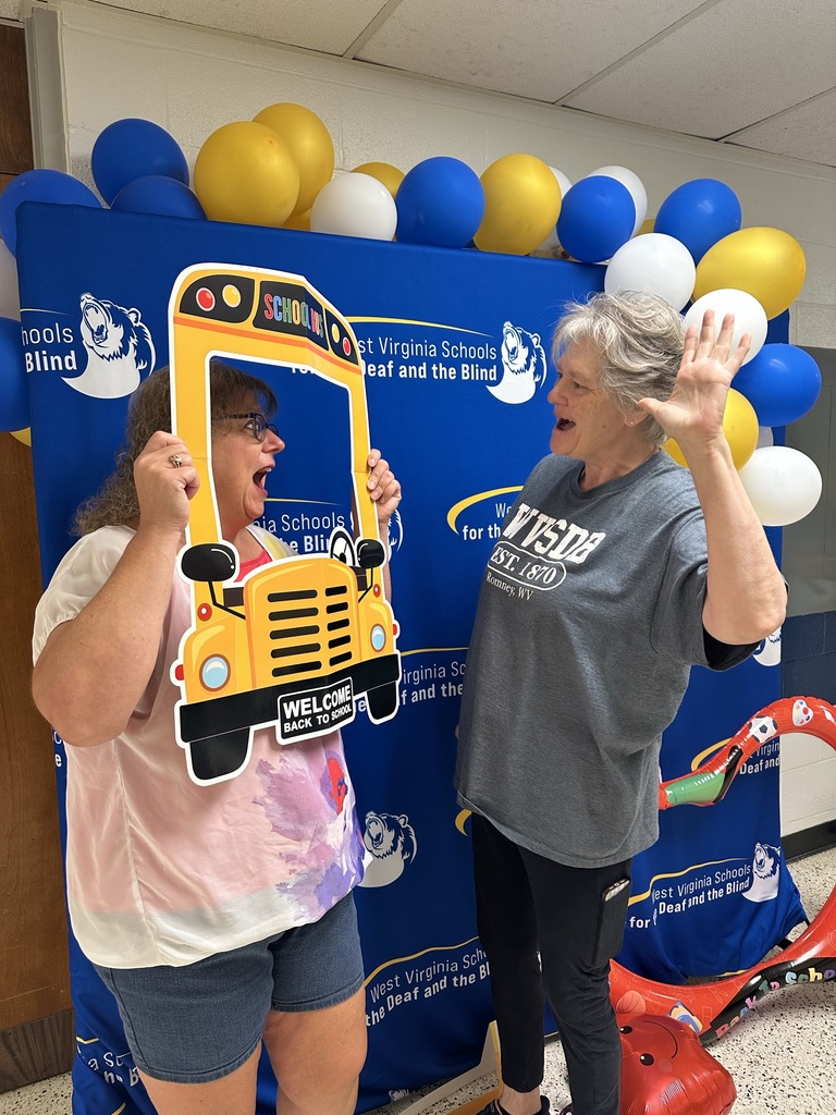 Two female aides make silly faces to one another as they participate in the photobooth activity in front of a blue backdrop.