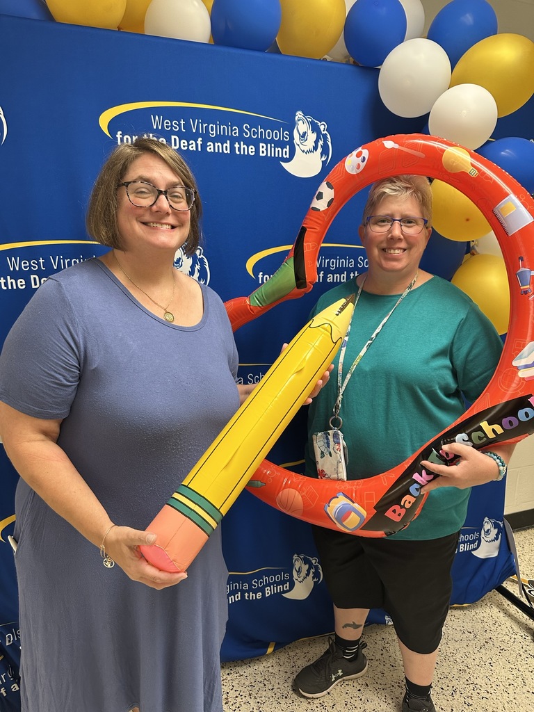 Two female teachers pose for a photo in front of a blue backdrop. The teacher on the left is holding a large inflatable pencil, while the teacher on the right holds an inflatable apple.