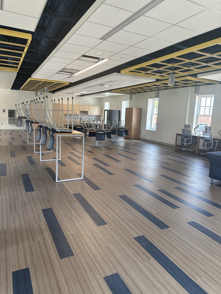An overview of the student center. Rectangular and circular-shaped tables with blue chairs fill the room sitting on top of gray and blue-colored flooring. In the background there is tan-colored cabinets and a vending machine.