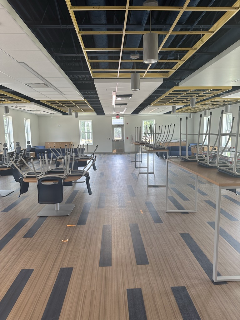 Tables and blue chairs line the center of the updated student center. There is gray and blue-colored flooring and blue and yellow-colored ceiling tile. There are windows around the perimeter of the student center.
