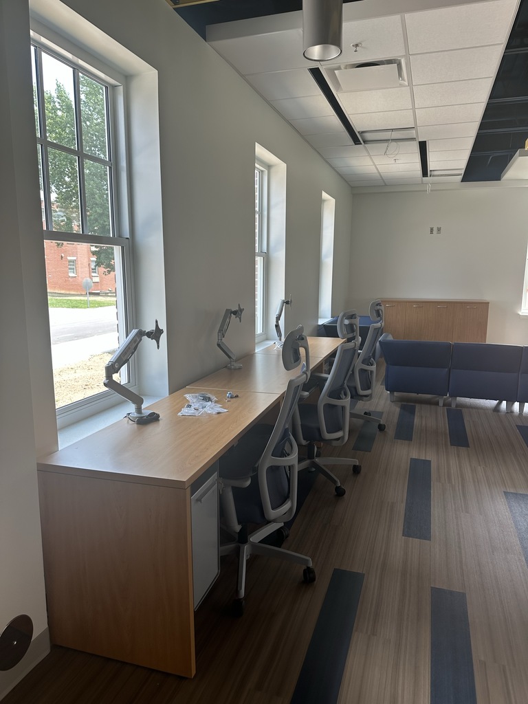 A large rectangular-shaped desk and three blue chairs sit up against a white wall and windows.